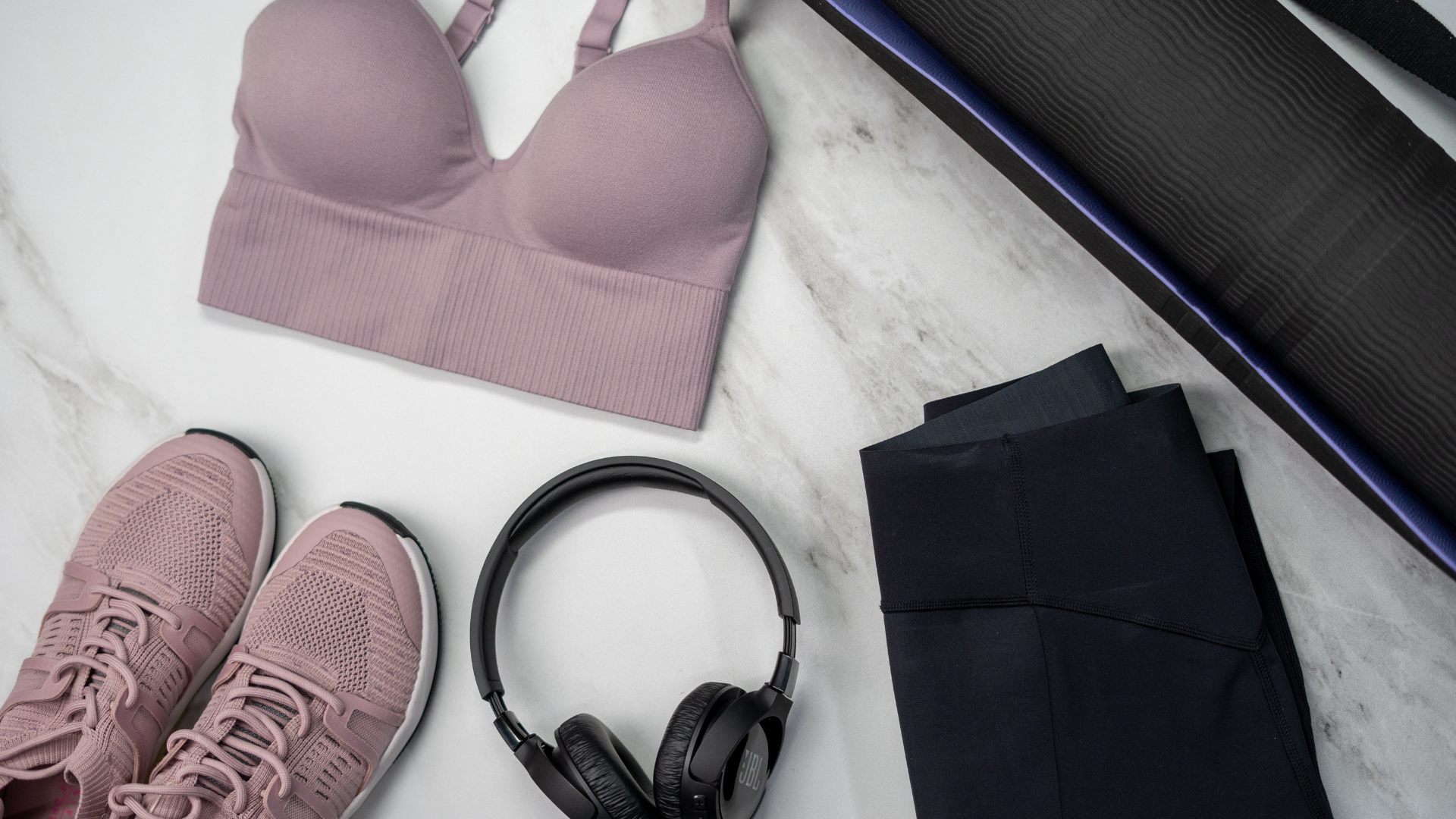 Un ensemble d'accessoires et de tenue yoga comprenant un haut de sport mauve, un legging noir, une paire de baskets assorties, un casque audio noir et un tapis de yoga noir et violet, posé sur un fond en marbre.